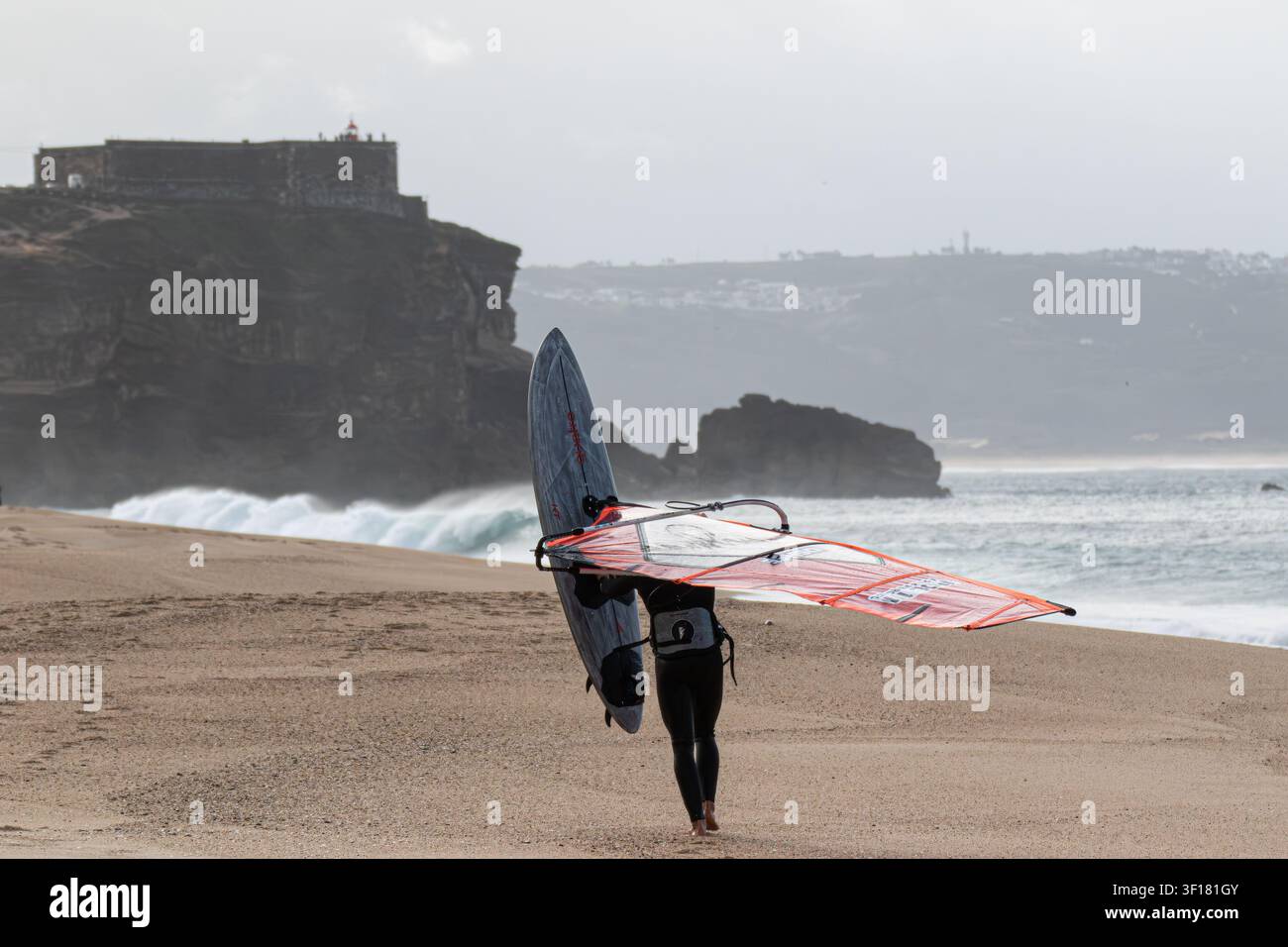 Windsurfeur portant l'équipement le long de la plage près du Forte de São Miguel Arcanjo à Nazaré, Portugal. Banque D'Images