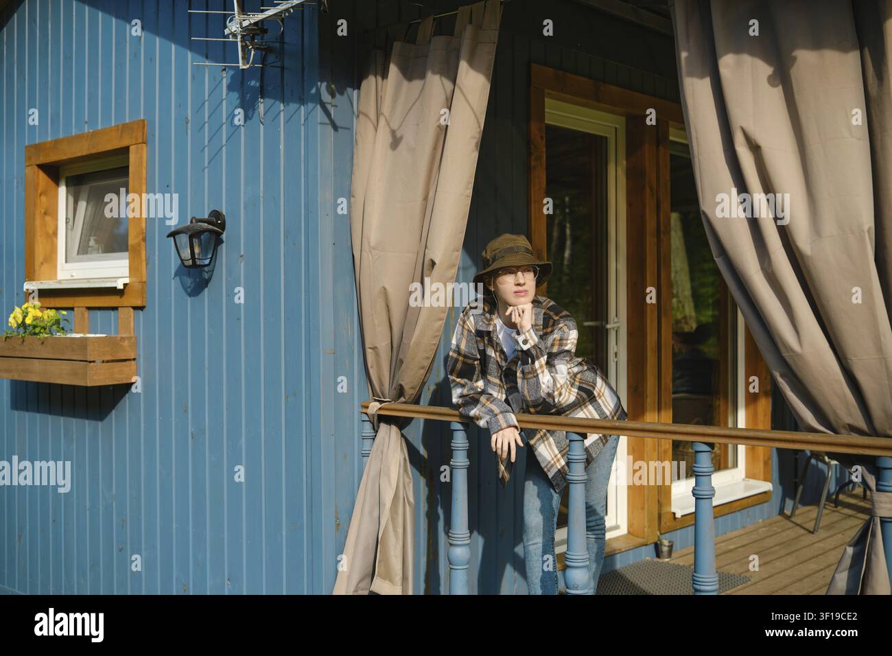 Jeune femme pensive appuyée sur la rambarde d'une cabane en bois bleue, portant un chapeau et une chemise à carreaux, regardant pensivement au loin. Bright sunlight shines on he Banque D'Images