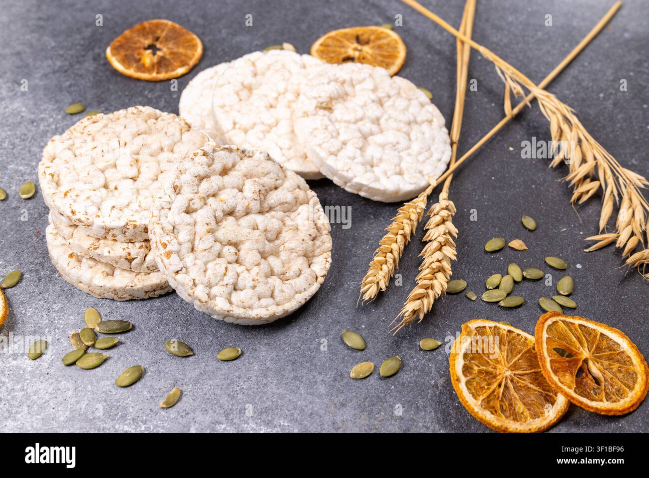 Plusieurs gâteaux de riz ronds avec des graines de courge, des rondelles d'orange séchées et des épis de blé éparpillés sur une surface gris foncé, représentant des options de collations saines Banque D'Images