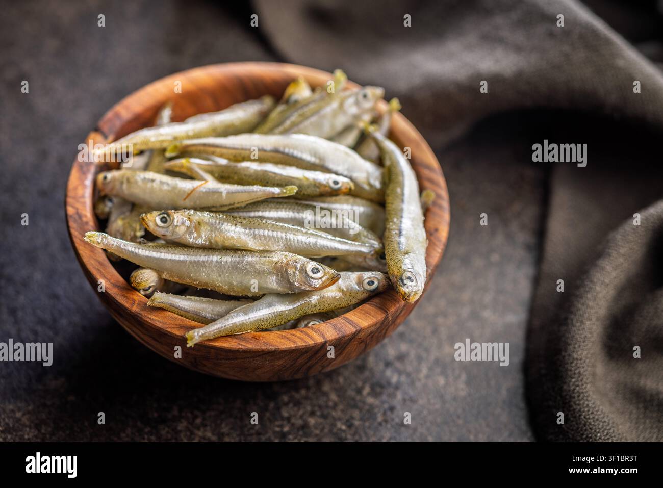 Petits poissons frais empilés dans un bol en bois sur une surface rustique, idéaux pour les recettes de fruits de mer, les entrées ou les plats méditerranéens. La texture naturelle et la présentation simple mettent en valeur la fraîcheur et l'utilisation culinaire. Banque D'Images
