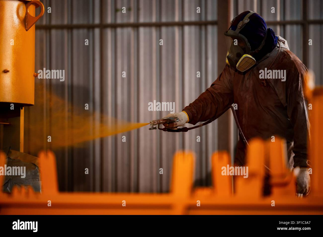 Ouvrier pulvérise de la peinture orange dans un environnement d'usine. Portant un casque et un masque à gaz, la personne se concentre sur la tâche. Structures métalliques et plaques ondulées w Banque D'Images
