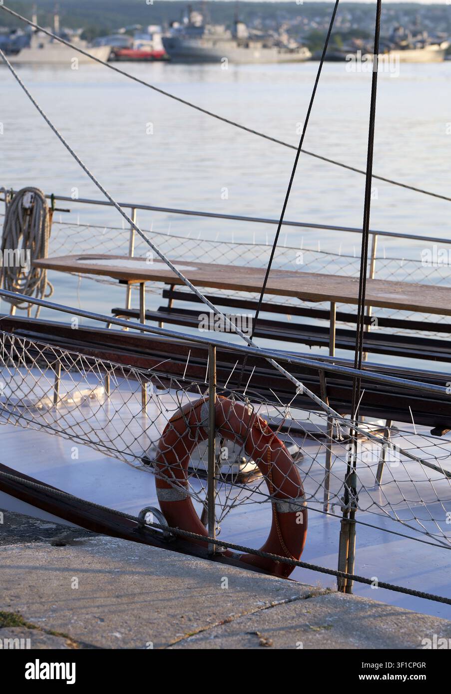 Pont de bateau à voile avec un anneau de sauvetage orange visible, un filet de protection, des cordages enroulés et une table en bois, avec d'autres navires et une côte dans le port Banque D'Images