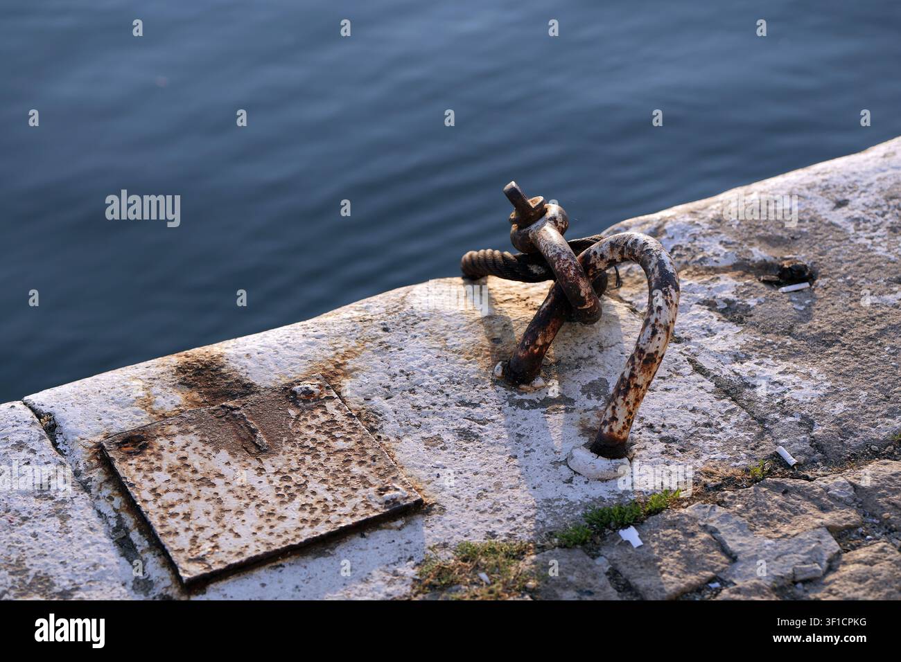 Bordure de quai en béton patiné comportant un anneau d'amarrage en métal rouillé et une plaque boulonnée, s'étendant au-dessus de l'eau calme et bleu profond de l'océan Banque D'Images