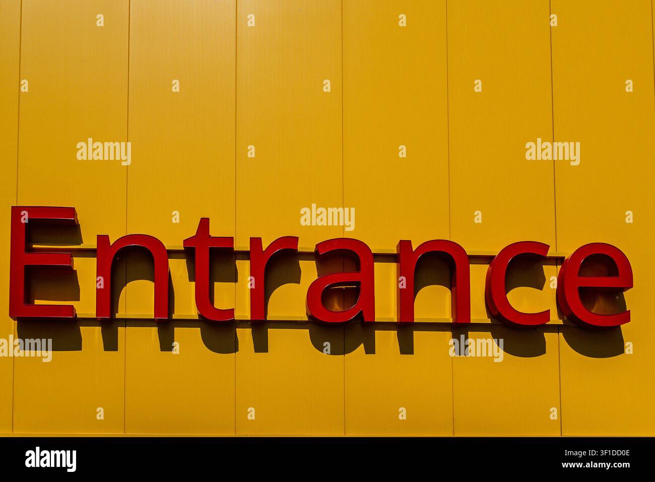 Un panneau 3D rouge "Entrée" monté sur une façade jaune vif texturée en Irlande, projetant des ombres nettes sous un soleil éclatant. Banque D'Images