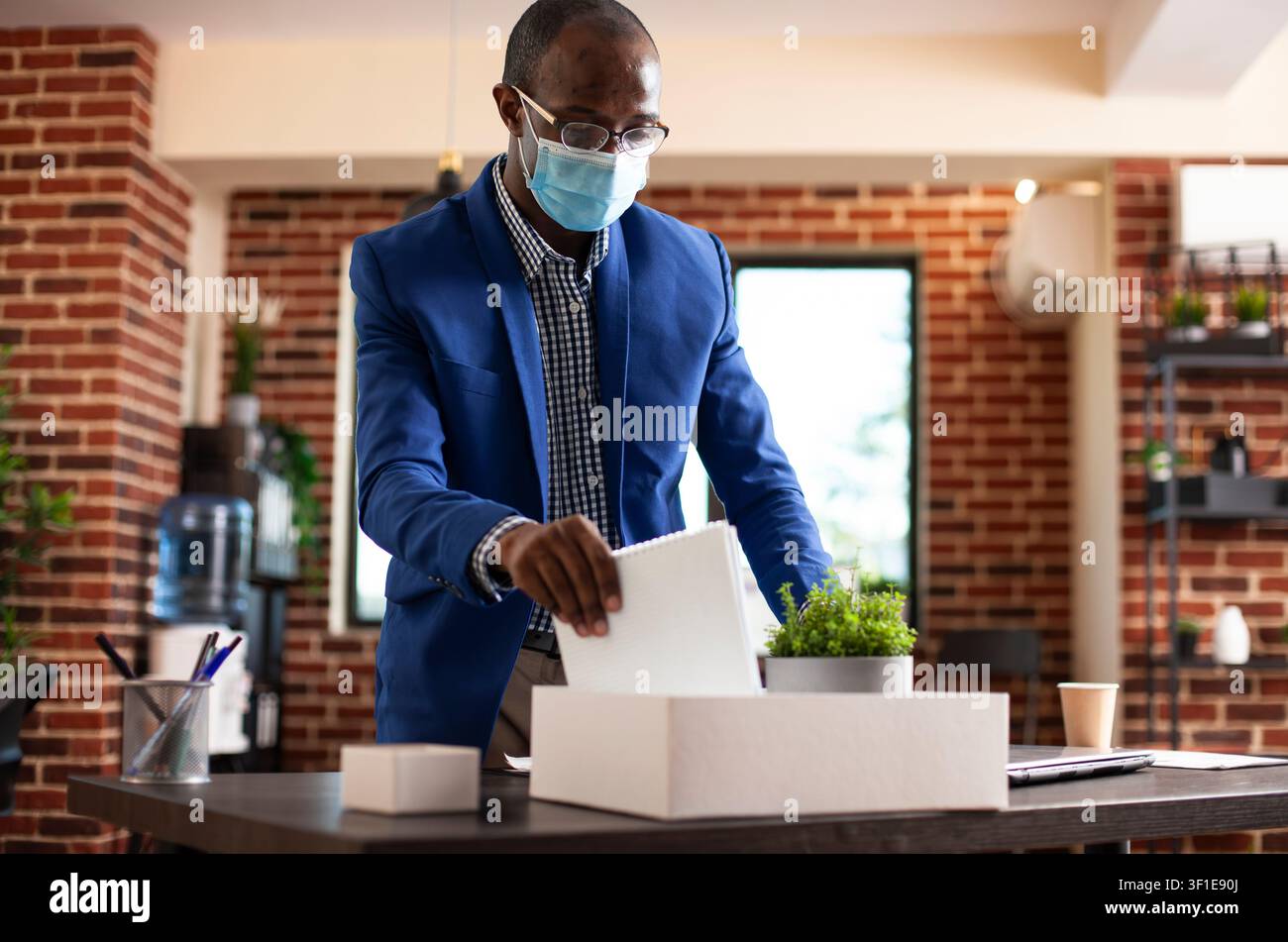 Gestionnaire organisant ses affaires sur le bureau, mettant en avant un gestionnaire masculin retirant un cahier d'un carton blanc et rangeant son bureau, homme noir en tenue de travail debout dans une pièce aux murs de briques. Banque D'Images