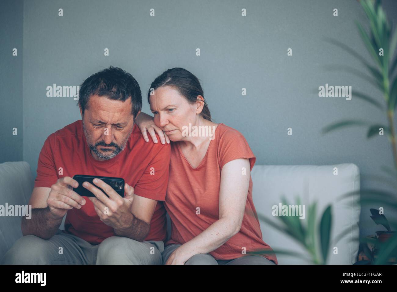 Un homme et une femme d'âge moyen sont assis côte à côte sur un canapé, lisant attentivement un message sur un smartphone. Leurs expressions traduisent de l'inquiétude, du soutien et une inquiétude partagée Banque D'Images