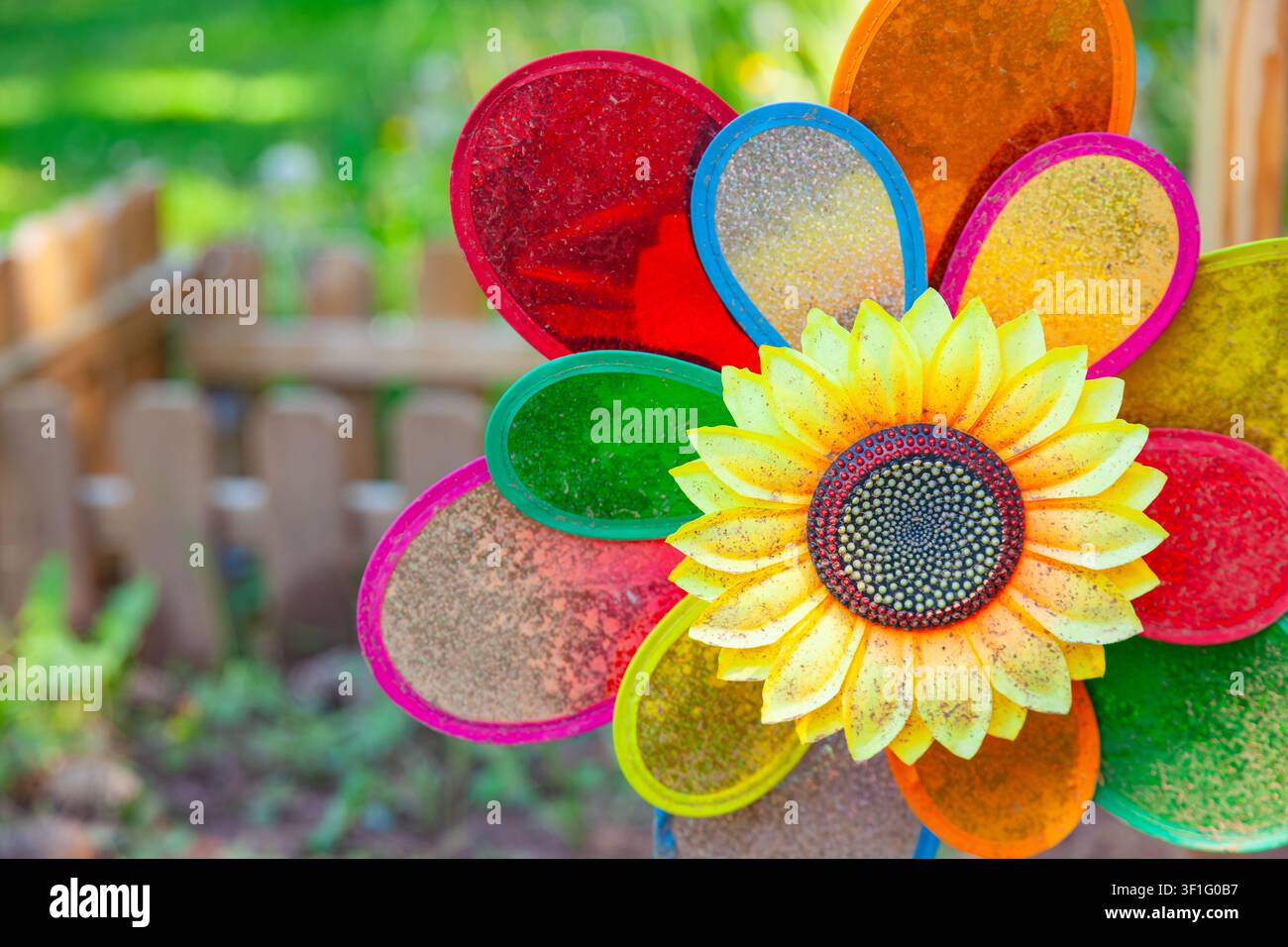 Moulin à vent coloré en forme de tournesol avec des pétales pailletés arc-en-ciel sur un fond flou de clôture de jardin en gros plan Banque D'Images