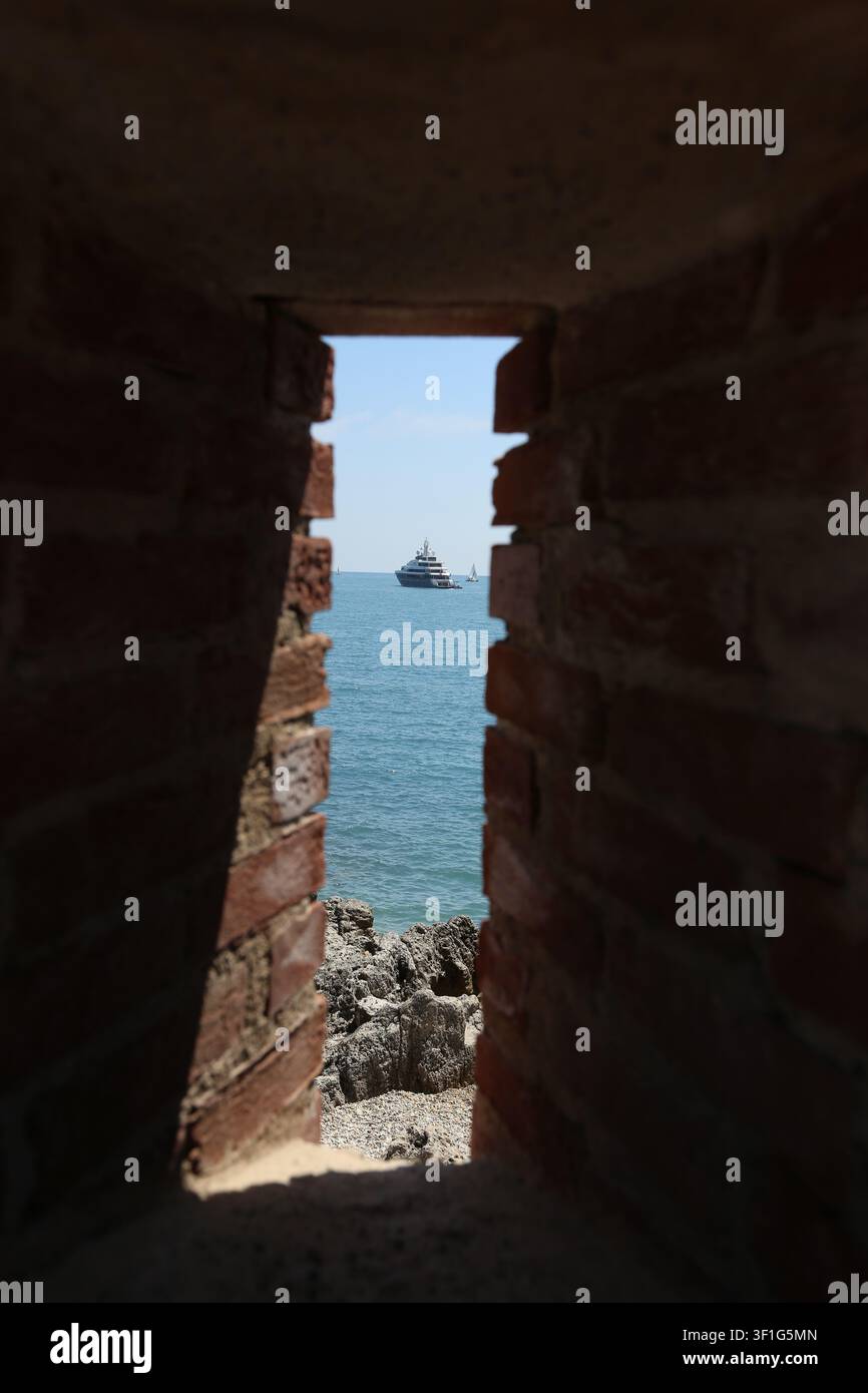 Plusieurs navires, dont un patrouilleur et un grand yacht, naviguent à l'horizon, vus à travers une arche de pierre érodée du Fort Carré à Antibes Banque D'Images