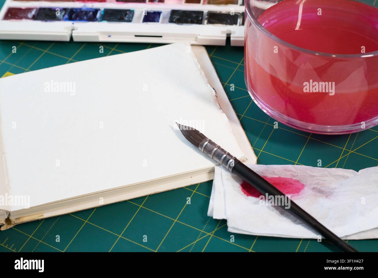 Page de papier aquarelle blanc propre sur le bureau. Pointe du pinceau sur un buvard près d'une tasse en verre rose contenant un liquide, vue aérienne des outils de table d'artiste Banque D'Images