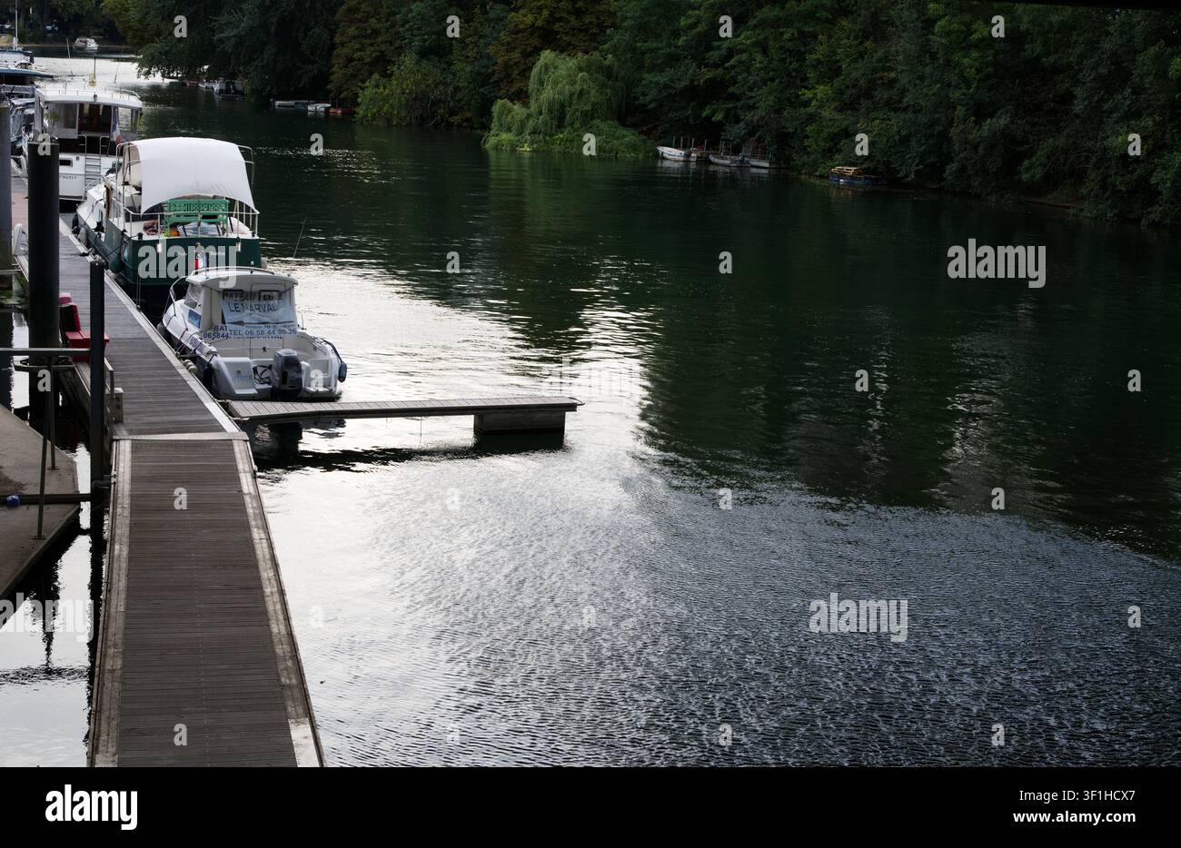 Scène agréable au bord de la rivière avec des bateaux de loisirs amarrés à une jetée en bois sur une rivière calme bordée d'arbres par une journée d'été paisible. Banque D'Images