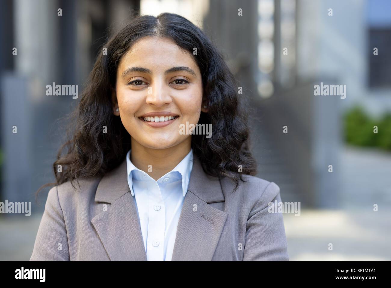Jeune femme d'affaires aux cheveux foncés et bouclés souriant largement, exprimant professionnalisme et succès tout en se tenant en plein air dans un environnement d'entreprise moderne Banque D'Images