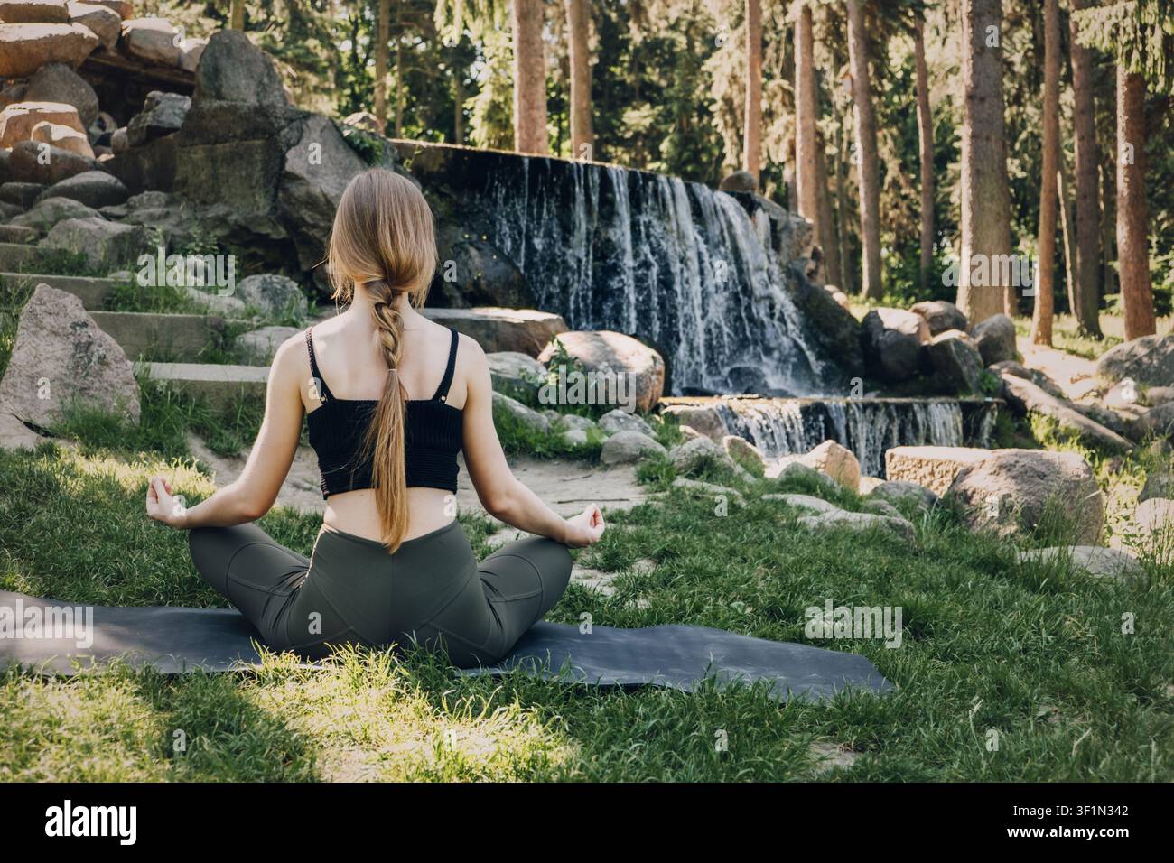 Jeune femme médite sur un tapis de yoga en plein air, cascade et pins en arrière-plan. Méditation de détox numérique, retraite de thérapie naturelle, spiritualité solitaire Banque D'Images