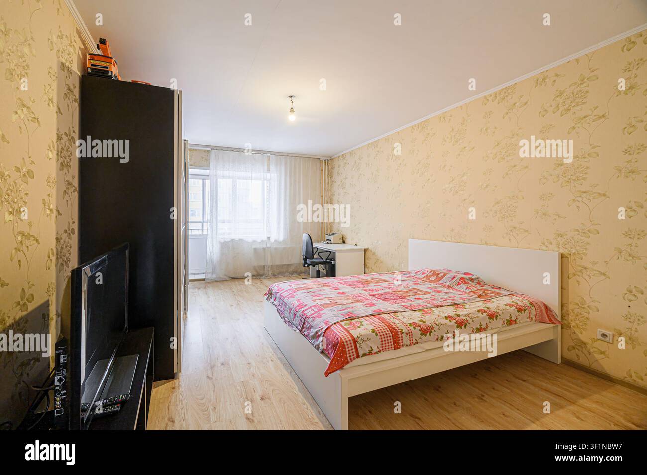chambre à coucher confortable avec un lit doté d'une couette fleurie, un bureau près de la fenêtre et une armoire noire. Les murs sont ornés d'un papier peint floral Banque D'Images