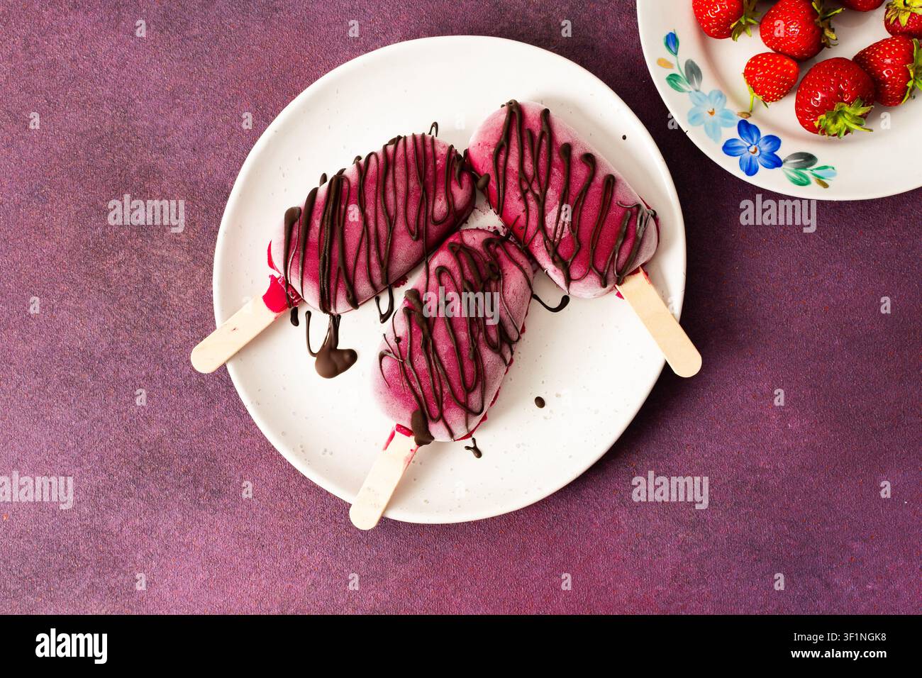 Trois popsicles à base de fraises, de bananes, de betteraves et de yaourt grec sont posées sur une assiette. Ils sont arrosés de chocolat et dégustés pendant un d ensoleillé Banque D'Images