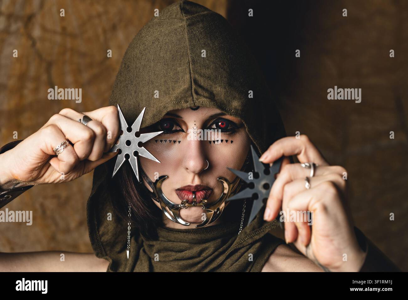 Portrait dramatique d'une femme dans un costume à capuche avec des lentilles de contact rouges, des bijoux de visage et des étoiles de lancer de style ninja, personnage de fantaisie sombre Hallowee Banque D'Images