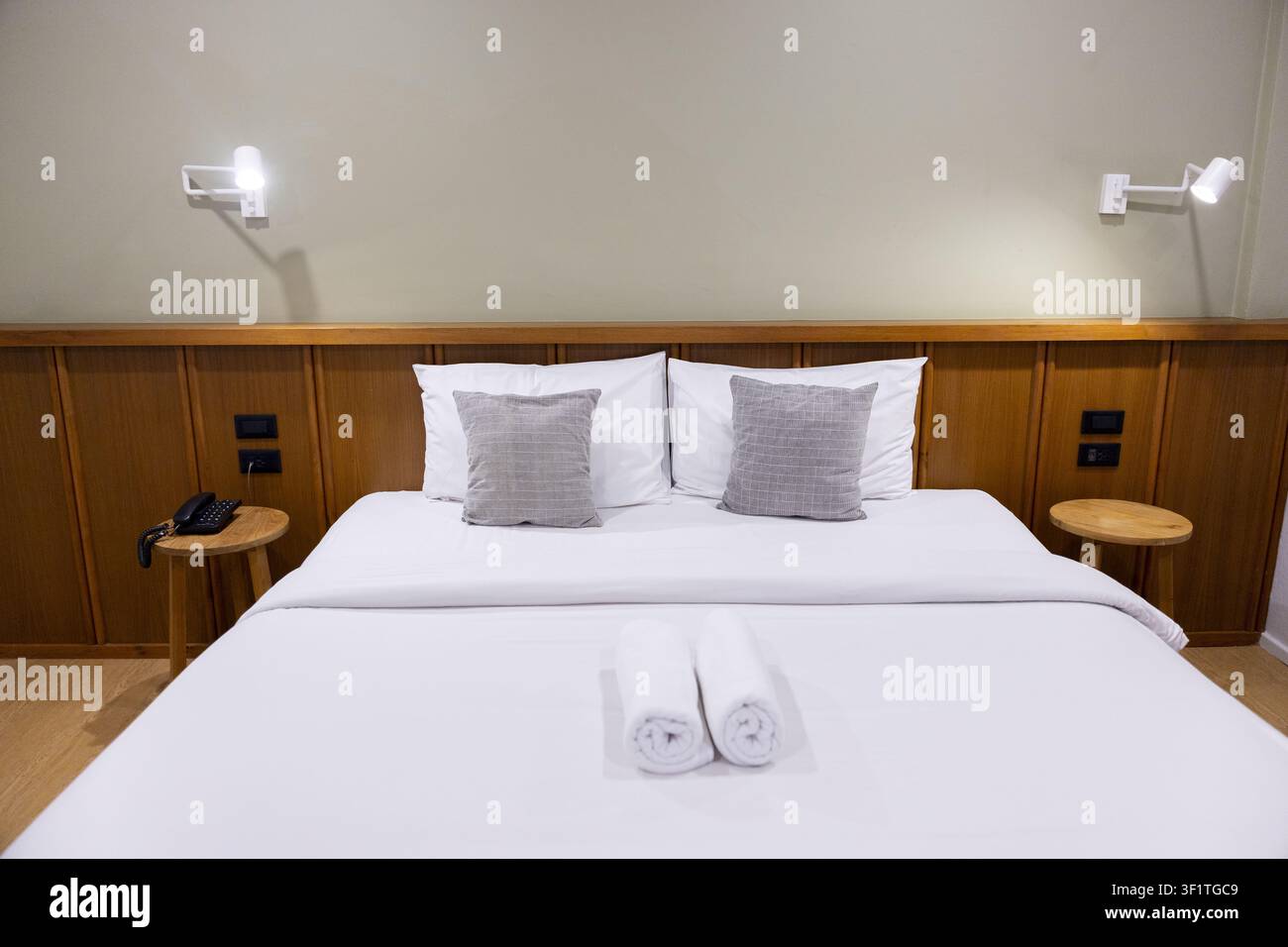 Un lit d'hôtel moderne avec des draps blancs et des oreillers gris soigneusement disposés sur une tête de lit en bois, des serviettes roulées placées sur la couette transmettant le calme et l'accueil Banque D'Images