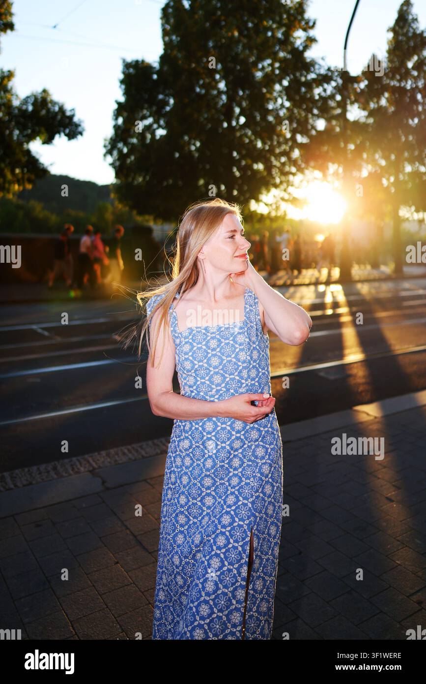 Belle jeune femme en robe d'été bleue marchant à l'heure dorée dans une ville européenne, souriant par-dessus son épaule dans la lumière chaude du coucher du soleil. Banque D'Images