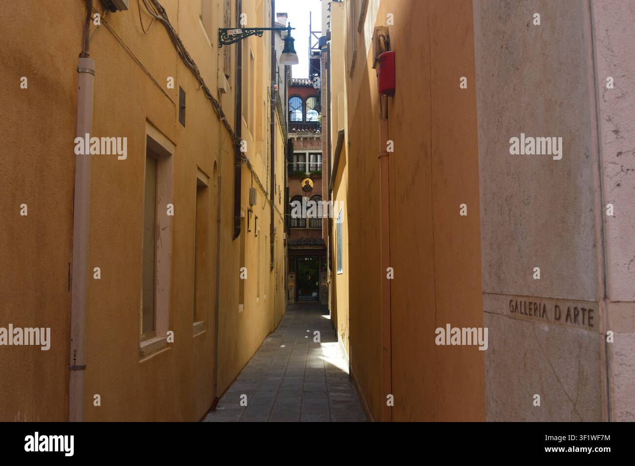 Ruelle vénitienne étroite et ensoleillée bordée de murs jaune chaud menant vers une enseigne de galerie d'art à Venise, Italie Banque D'Images