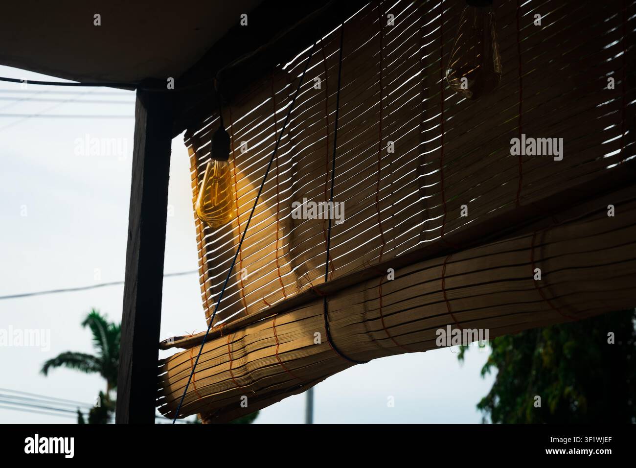 La lumière du soleil filtre à travers des stores en bambou partiellement roulés sur une véranda, créant des motifs de lumière et d'ombre naturels dans un cadre résidentiel tropical. Banque D'Images