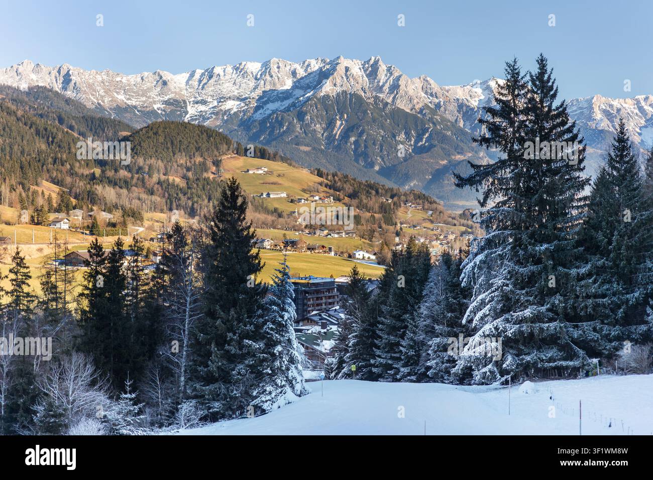 Une neige clairsemée recouvre les pentes alpines alors que les champs verts et le sol exposé contrastent avec les sommets enneigés sous un ciel d'hiver clair. La vallée montagneuse reflète la chaleur Banque D'Images