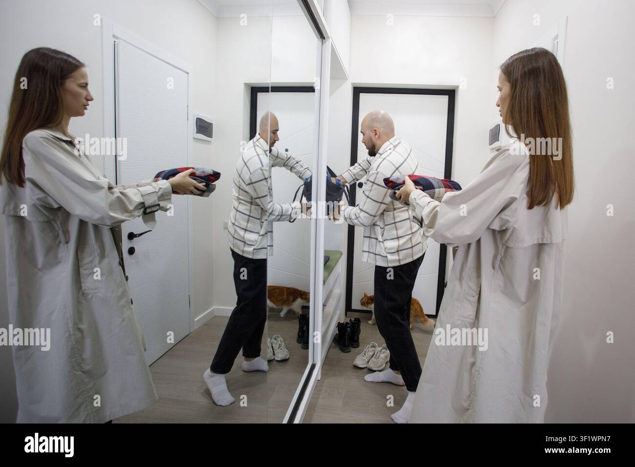 Une personne se tient devant un miroir en utilisant un sèche-cheveux. Une autre personne ajuste sa tenue à proximité. Un chat est assis au sol en arrière-plan. Le set Banque D'Images