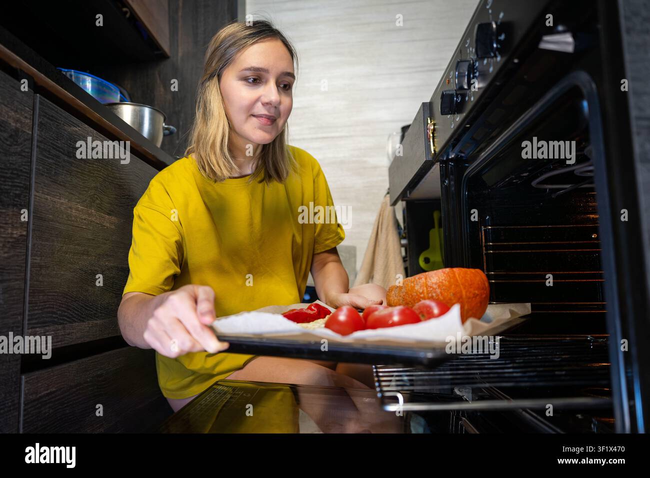 Jeune femme mettant une plaque de cuisson avec des légumes frais dans le four dans une cuisine moderne à la maison. Cuisine saine à domicile, préparation de repas, foo végétarien Banque D'Images