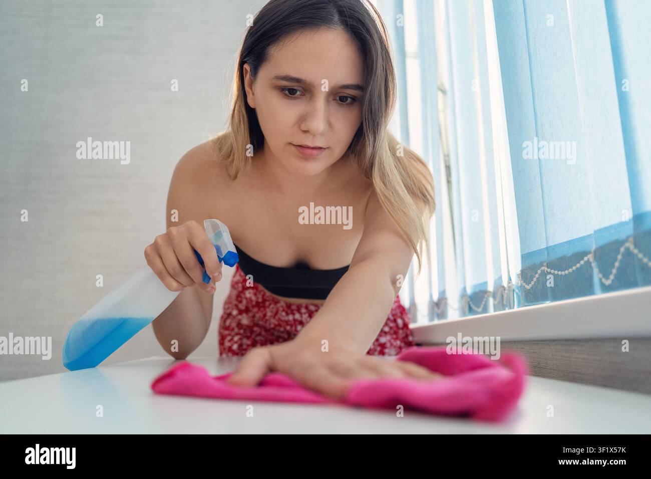 Femme essuyant une table blanche avec un chiffon rose tout en tenant une bouteille pulvérisatrice contenant un liquide bleu. Nettoyage domestique, tâches ménagères et hygiène des surfaces. Banque D'Images