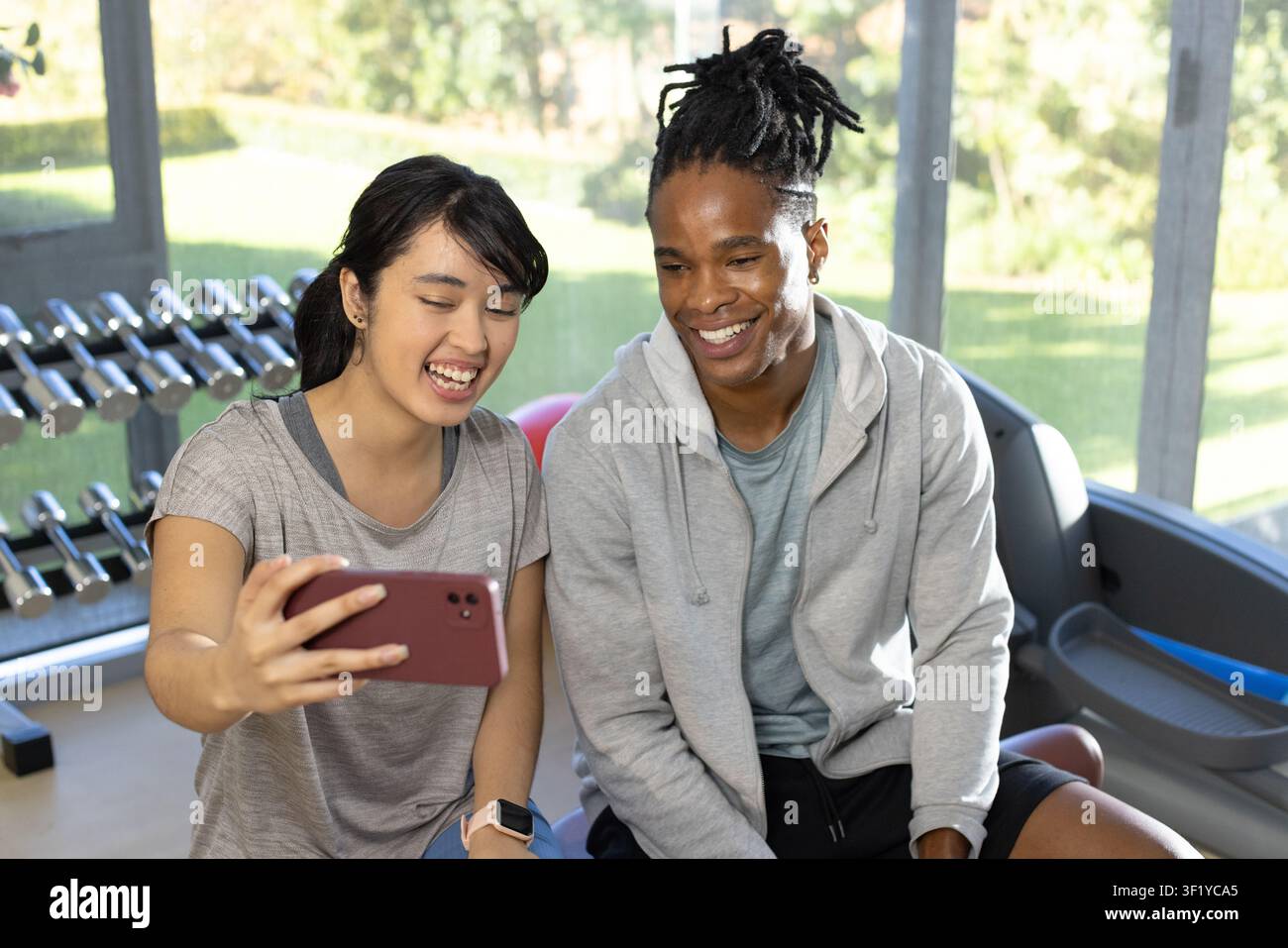 Couple divers en tenue de sport prenant un selfie sur un banc au gymnase avec un smartphone, étagère à haltères Banque D'Images