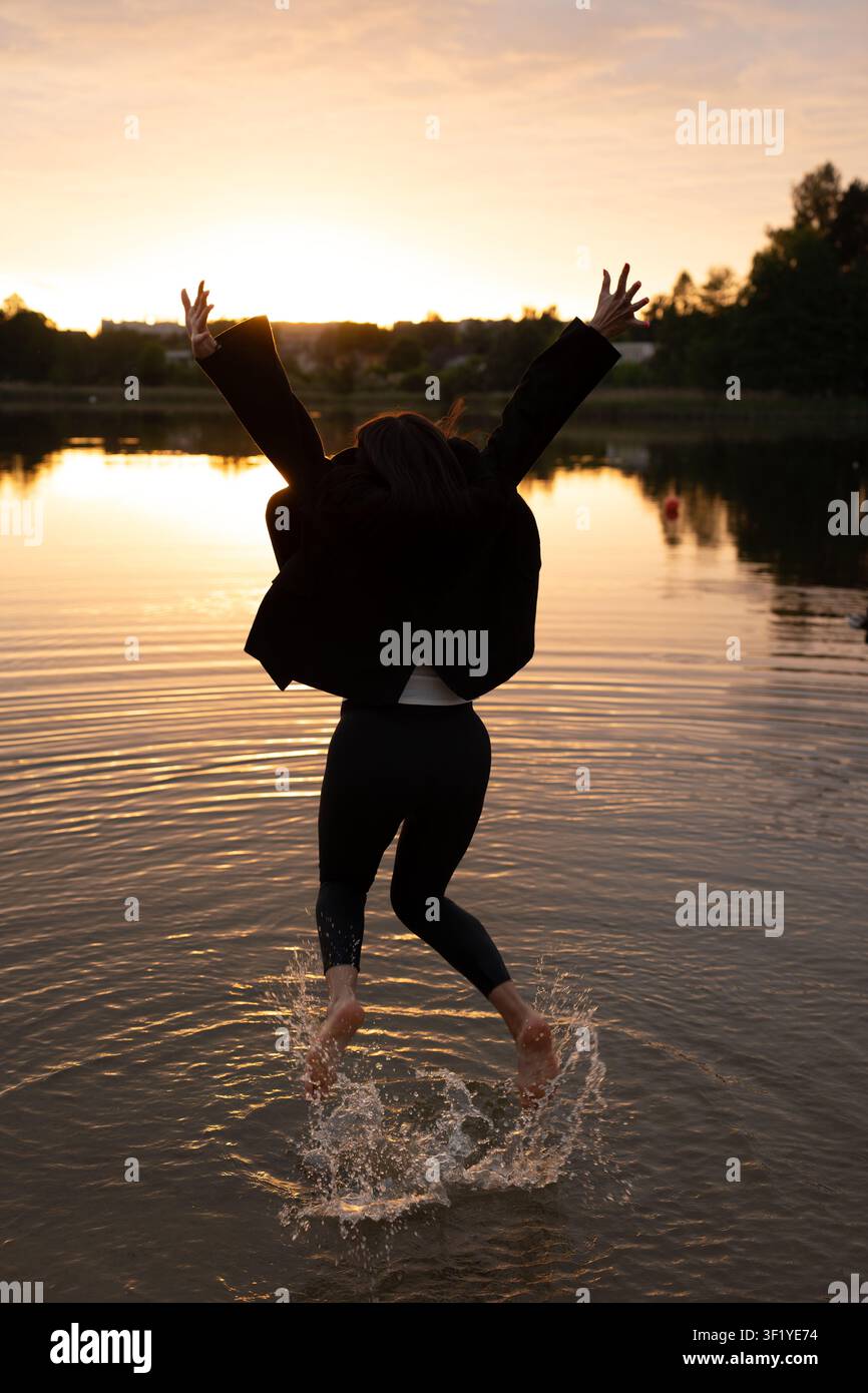 personne en silhouette sautant dans un lac peu profond au coucher du soleil doré, les bras levés dans un mouvement exubérant, réflexions orange chaudes et eau ondulante, chaussure sombre Banque D'Images