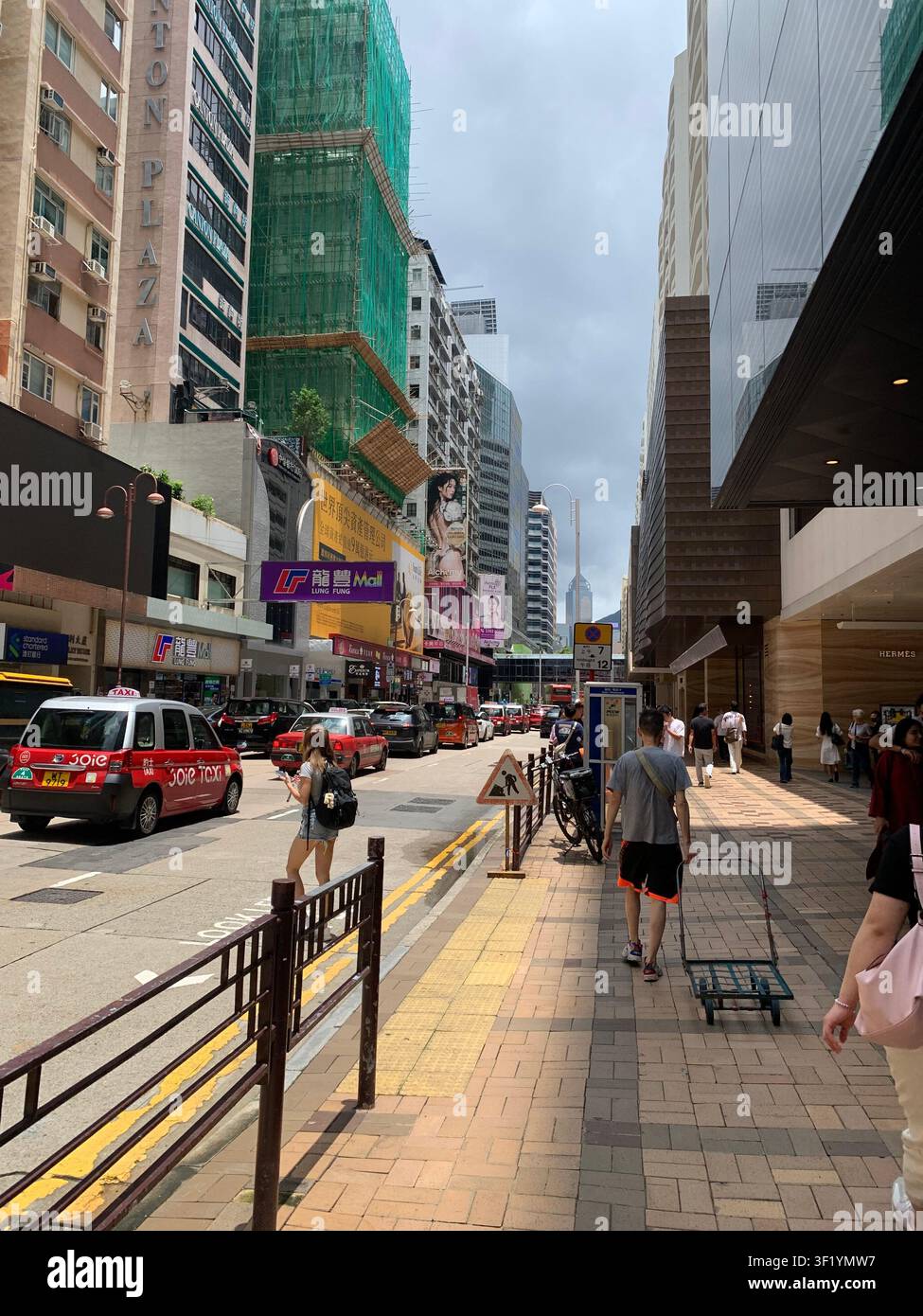 Une scène de rue animée à Hong Kong, mettant en vedette des taxis rouges emblématiques, des acheteurs et des immeubles commerciaux sous un ciel nuageux - Image de stock capturée avec un smartphone