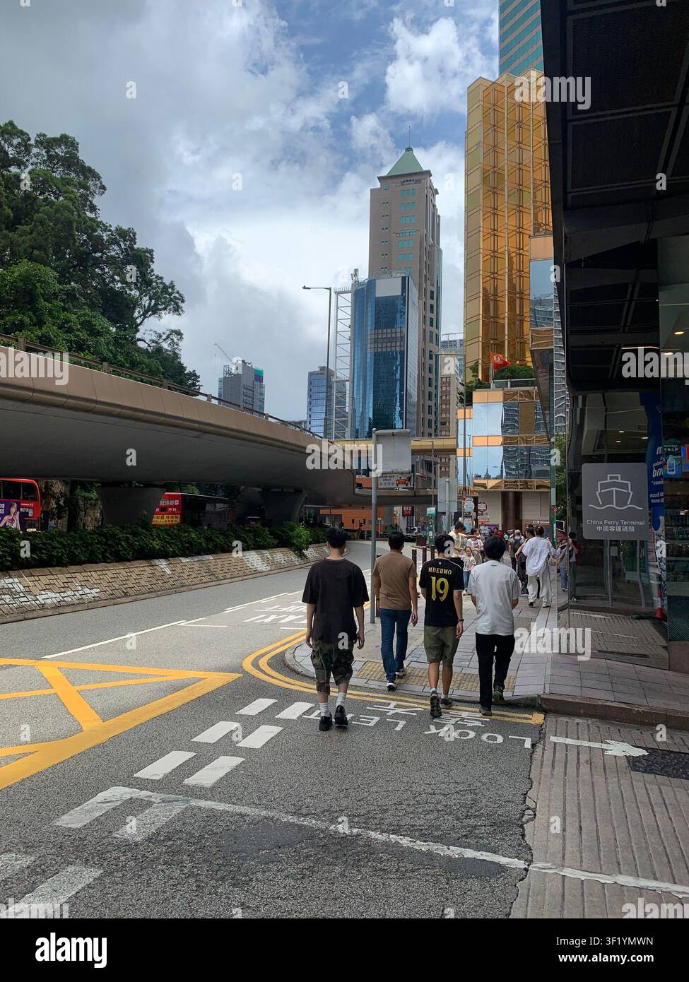 Des piétons marchent le long d'une rue de la ville de Hong Kong près du China Ferry Terminal, avec des gratte-ciel modernes et une passerelle surélevée en arrière-plan. - Image de stock capturée avec un smartphone