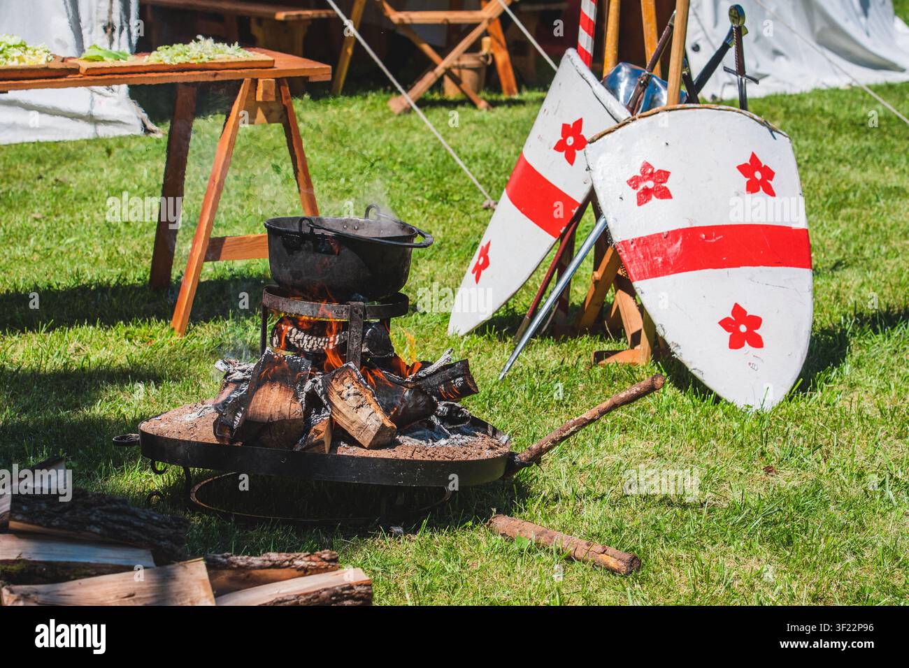 Un campement historique en plein air présente un chaudron cuisant sur une fosse à feu à côté de deux boucliers blancs et rouges lors d'une foire sur le thème médiéval Banque D'Images