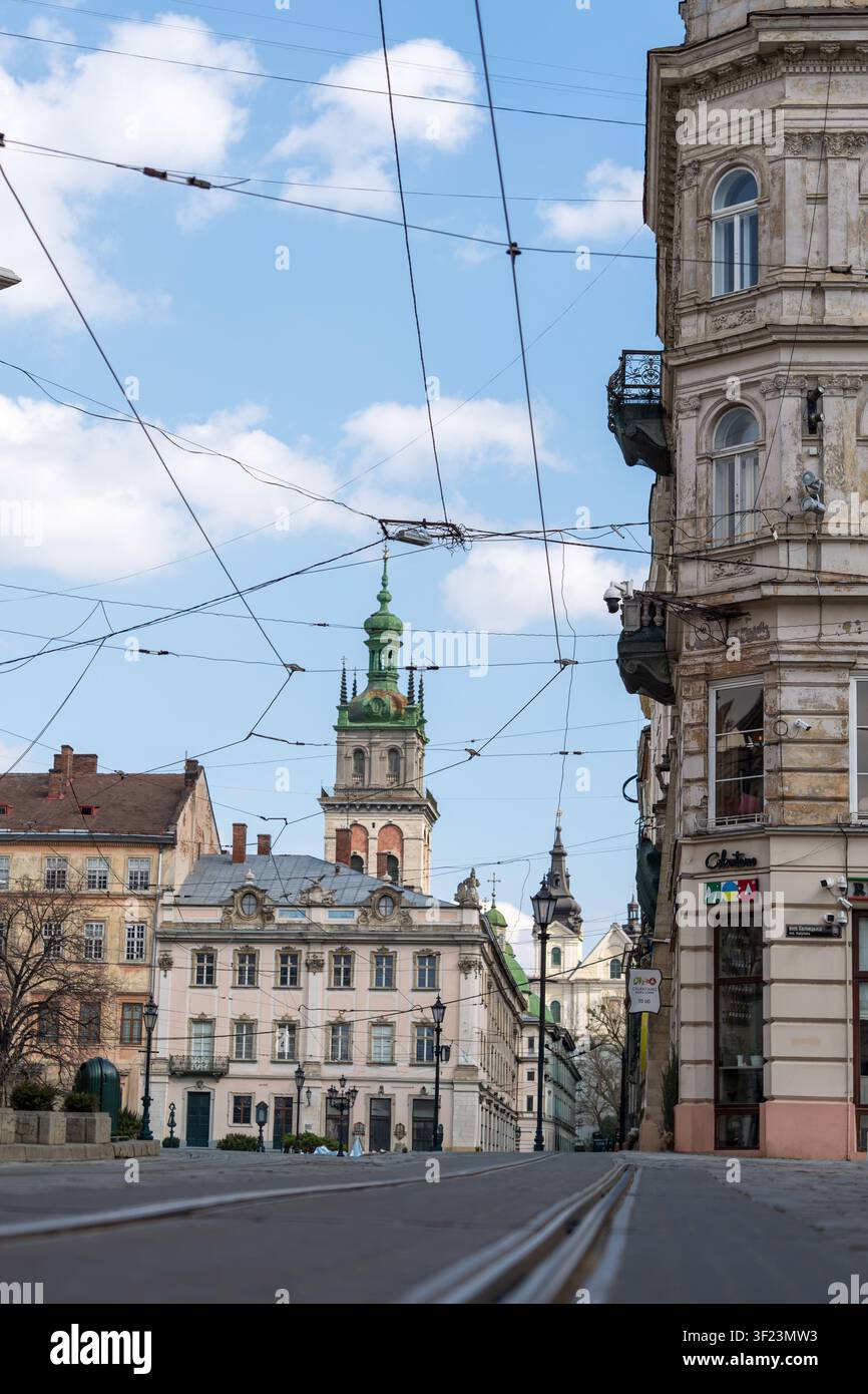 Une vue de rue à Lviv montre des bâtiments d'architecture historique. Le ciel est bleu avec quelques nuages. Des fils électriques traversent au-dessus de la rue. Les gens marchent Banque D'Images