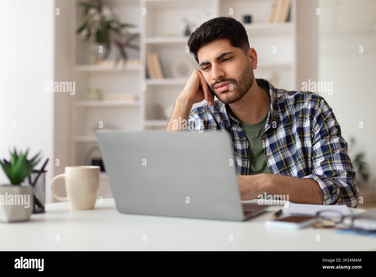 Jeune homme concentré en chemise à carreaux travaillant sur un ordinateur portable au bureau à domicile, concept commercial Banque D'Images