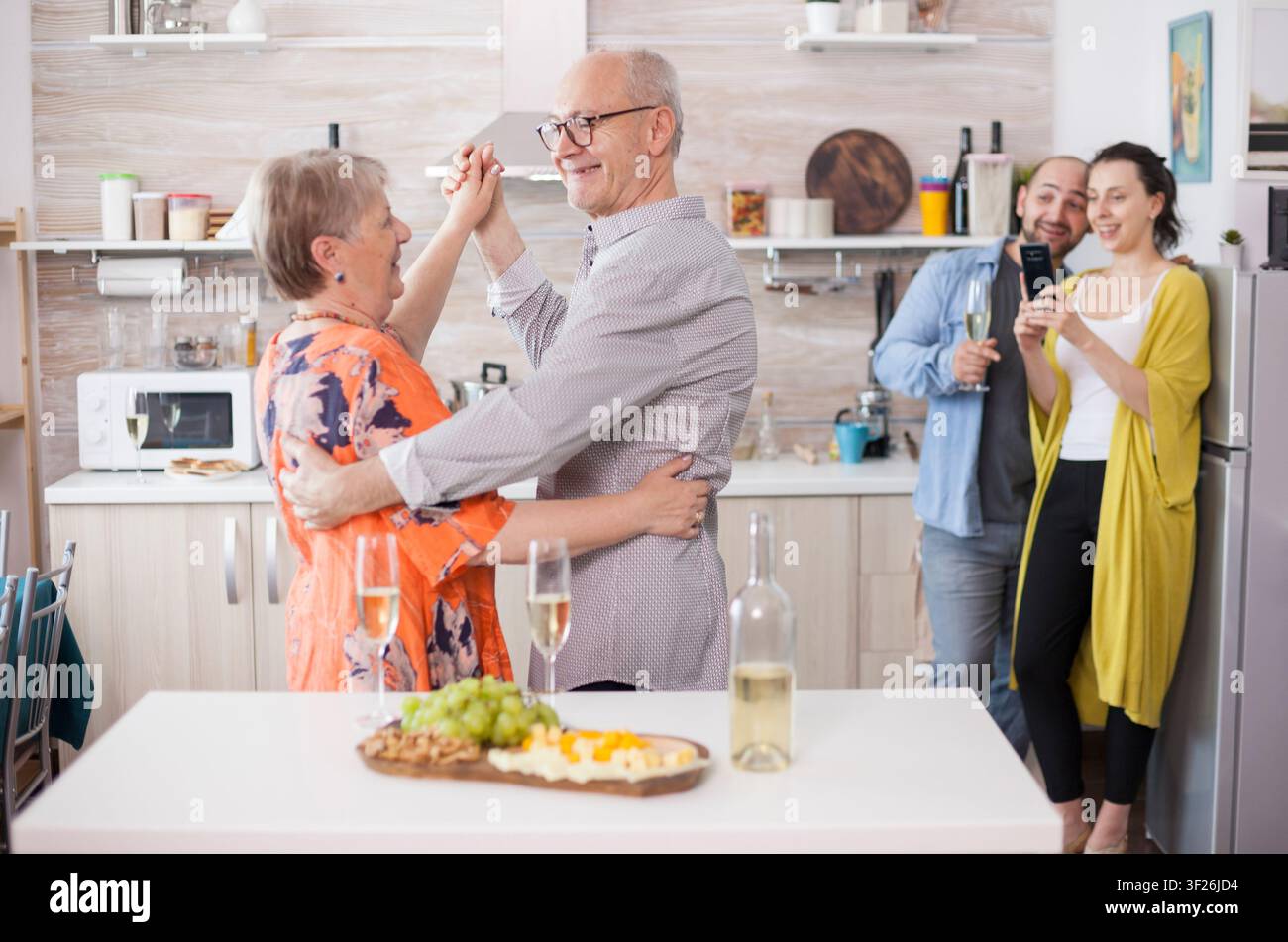 Un couple de personnes âgées joyeux danse dans la cuisine lors d'une célébration familiale, avec divers fromages sur une planche en bois et une femme qui prend des photos. Banque D'Images