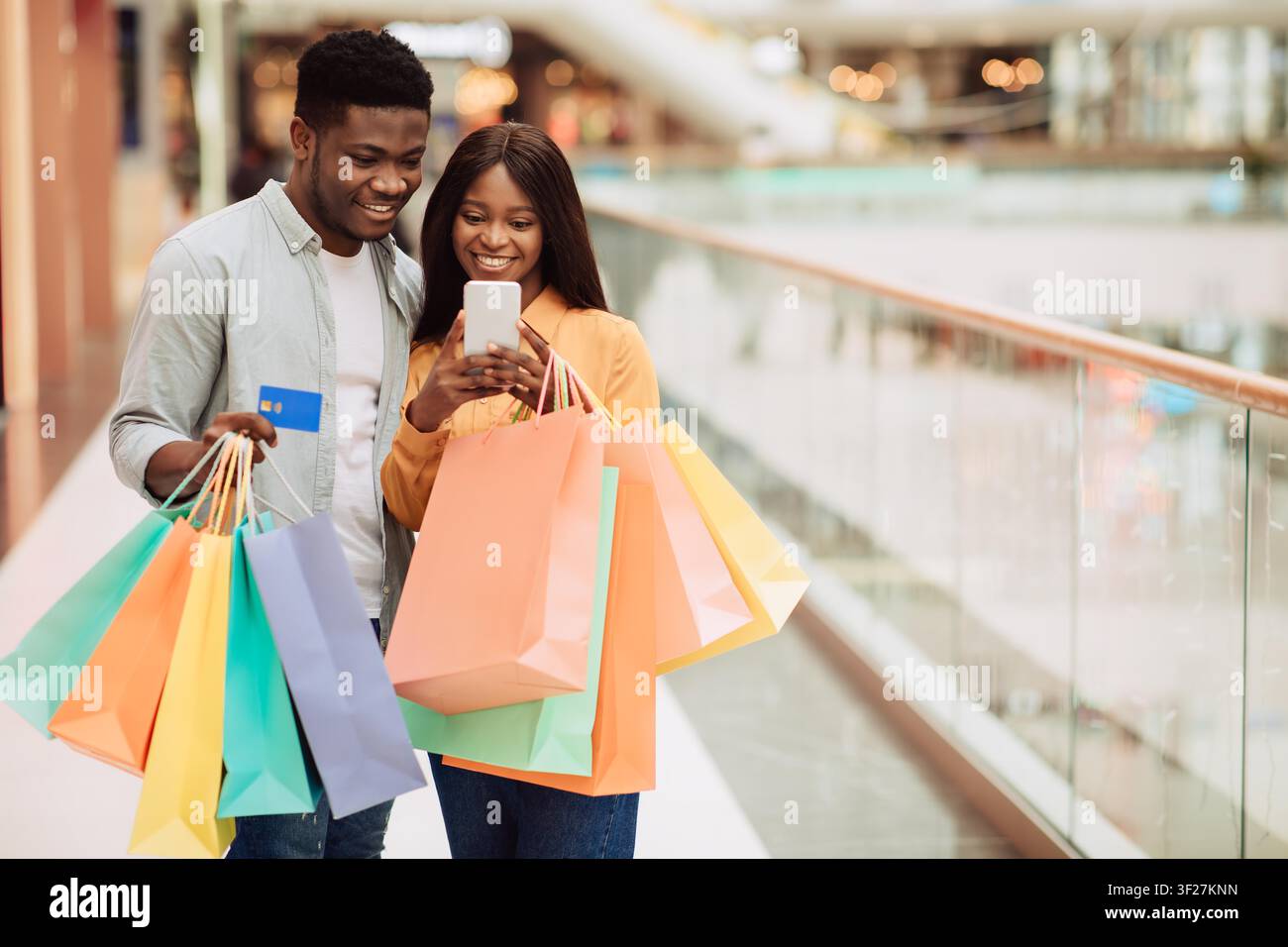 Couple noir heureux faisant ses courses ensemble au centre commercial, regardant leur smartphone avec des sacs de courses Banque D'Images