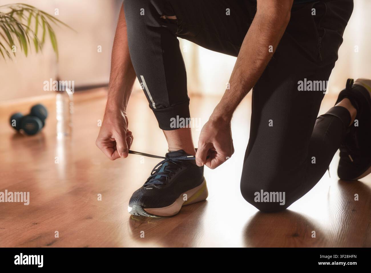 Homme attachant une baskets dans une pièce au sol en parquet, vêtu de vêtements de sport, avec des haltères et une bouteille d'eau en arrière-plan. Banque D'Images