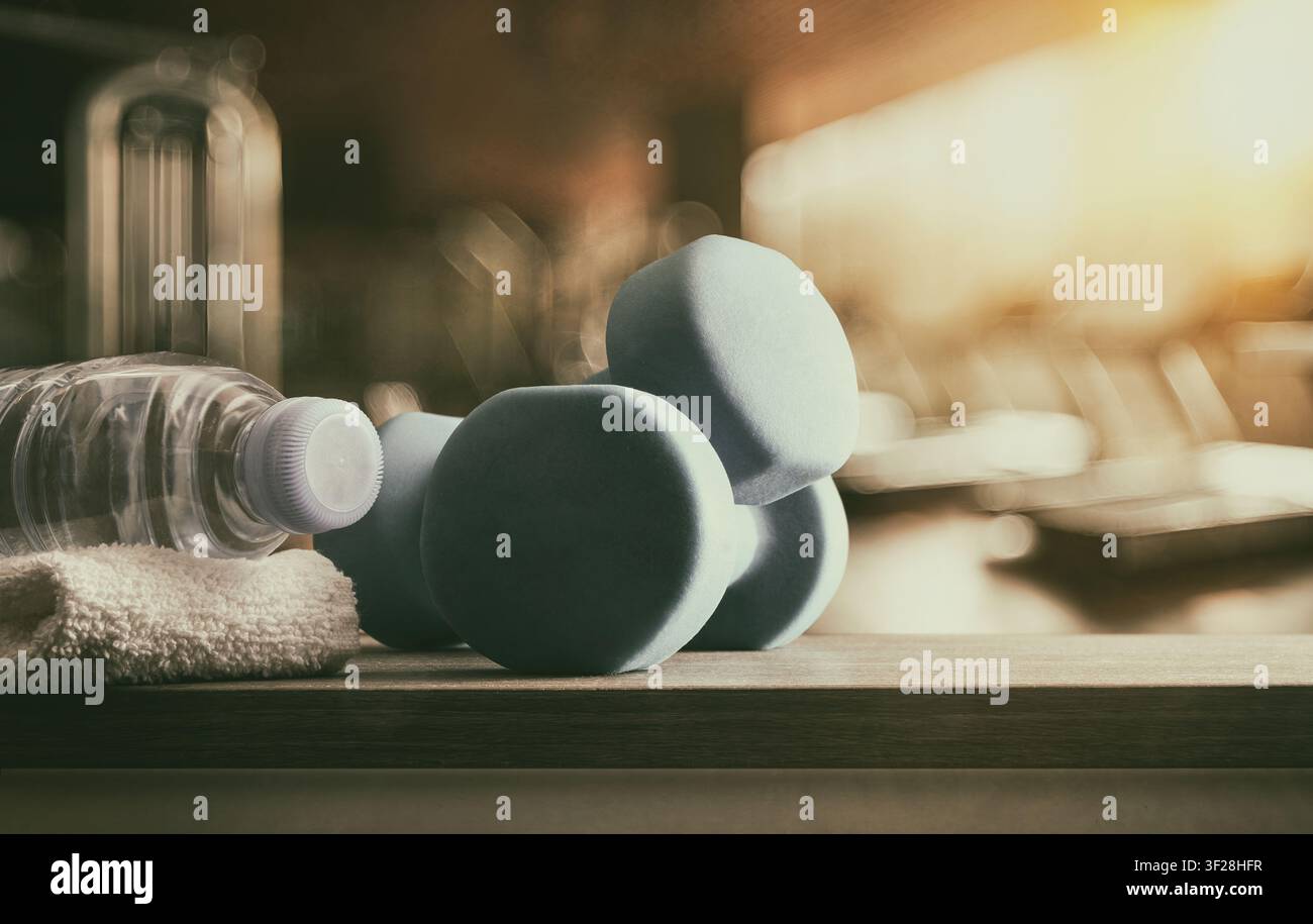 Haltères en mousse bleue, une bouteille d'eau et une serviette sur un banc de gym avec des équipements de fitness flous, une lumière chaude et une atmosphère d'entraînement motivante. Banque D'Images