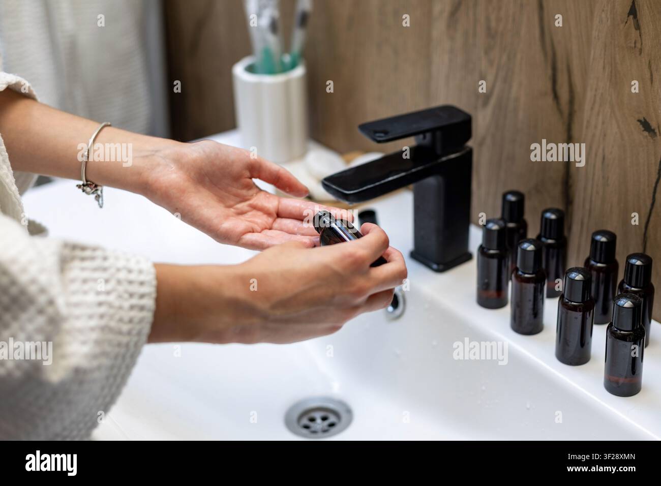 Femme manipulant des bouteilles ambrées au lavabo de la salle de bain. Une femme manipule une petite bouteille ambrée à côté de plusieurs contenants assortis sur un lavabo avec un noir Banque D'Images