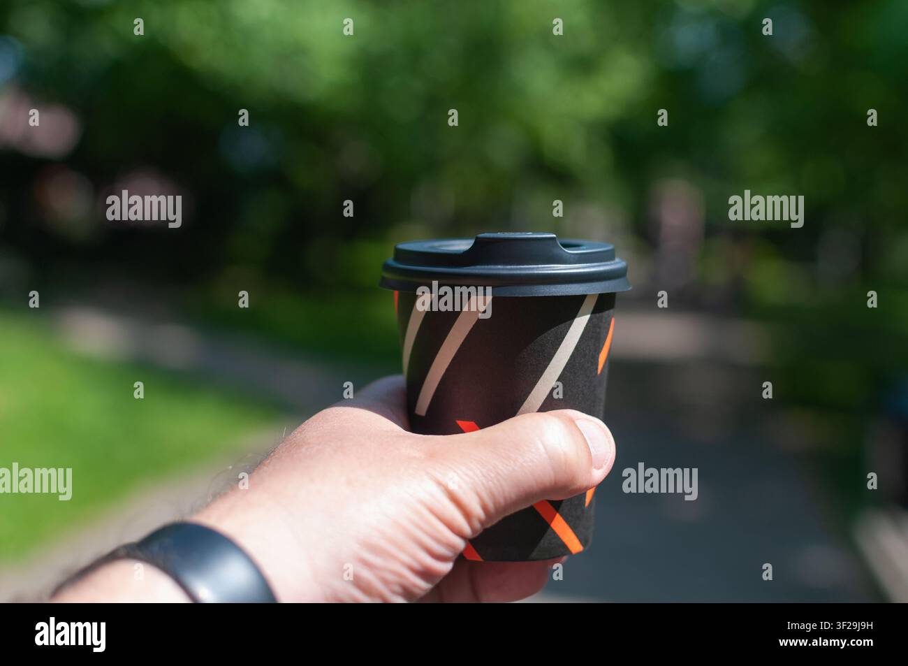 Une main d'homme tient une tasse à café en papier noir à emporter avec un couvercle noir tout en marchant à l'extérieur dans un parc vert ensoleillé Banque D'Images