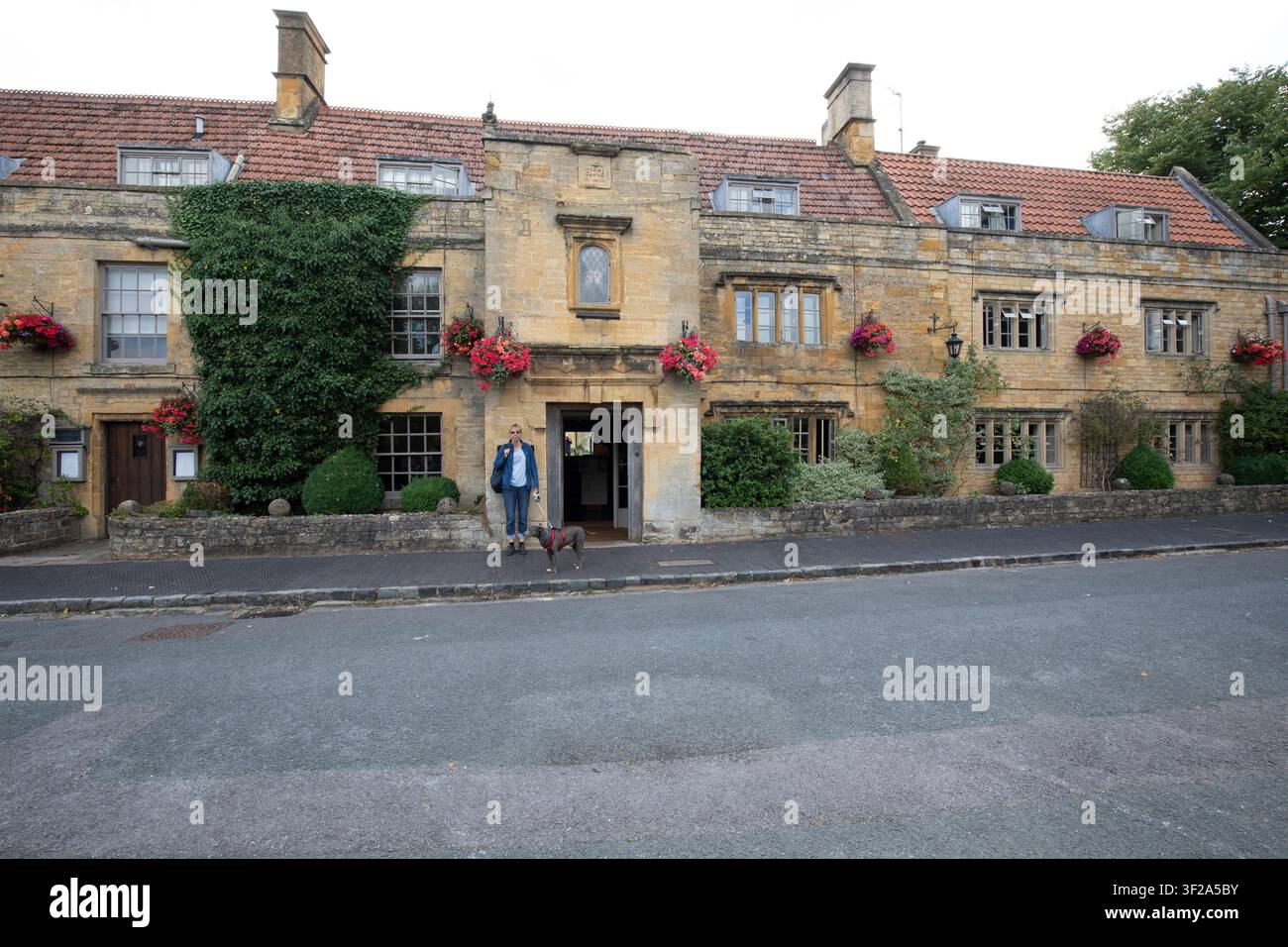 Une femme debout avec son chien de compagnie devant l'entrée du The Manor House Hotel sur la High Street à Moreton-in-Marsh, Gloucestershire, Royaume-Uni. Banque D'Images