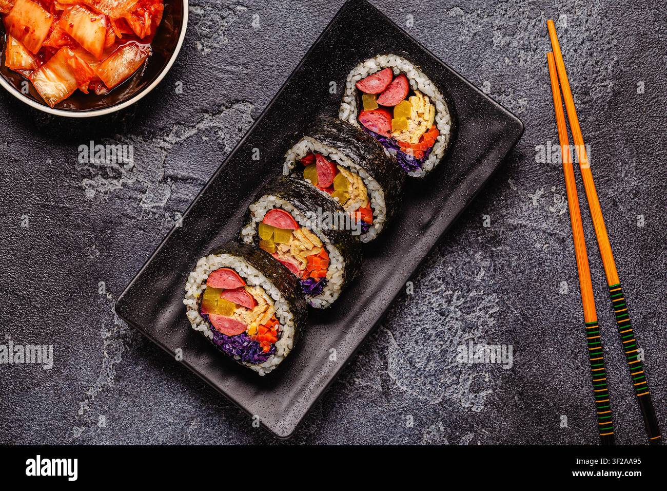 Une assiette noire remplie de kimbap (ou gimbap) aux garnitures colorées. Les ingrédients comprennent des saucisses, des cornichons, des œufs et des légumes. Banque D'Images