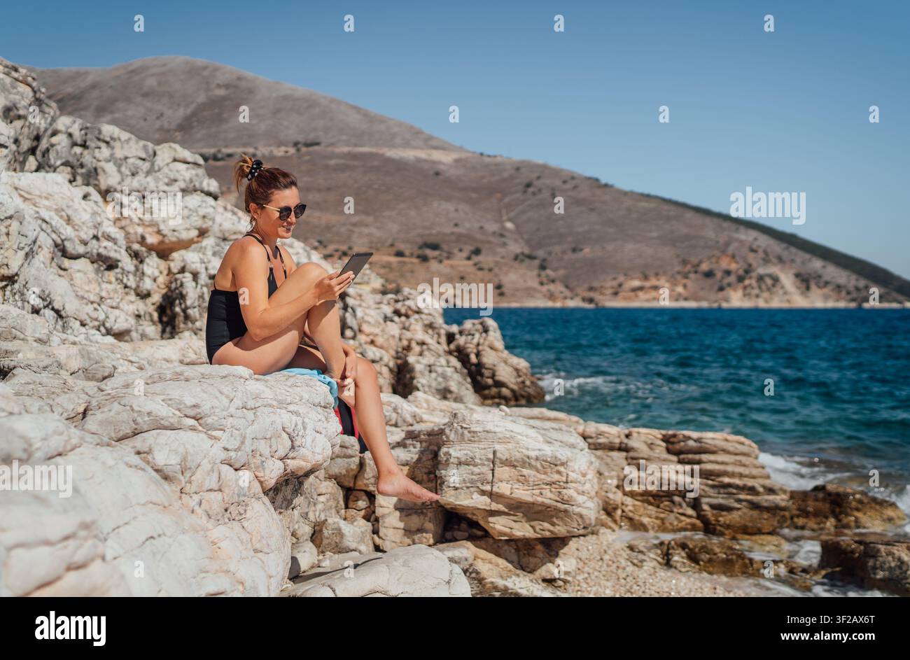 Jeune femme en maillot de bain noir assise sur la côte rocheuse de Céphalonie, Grèce, lisant un livre électronique, femme voyageuse heureuse se relaxant au bord de la mer Ionienne bleue avec sceni Banque D'Images