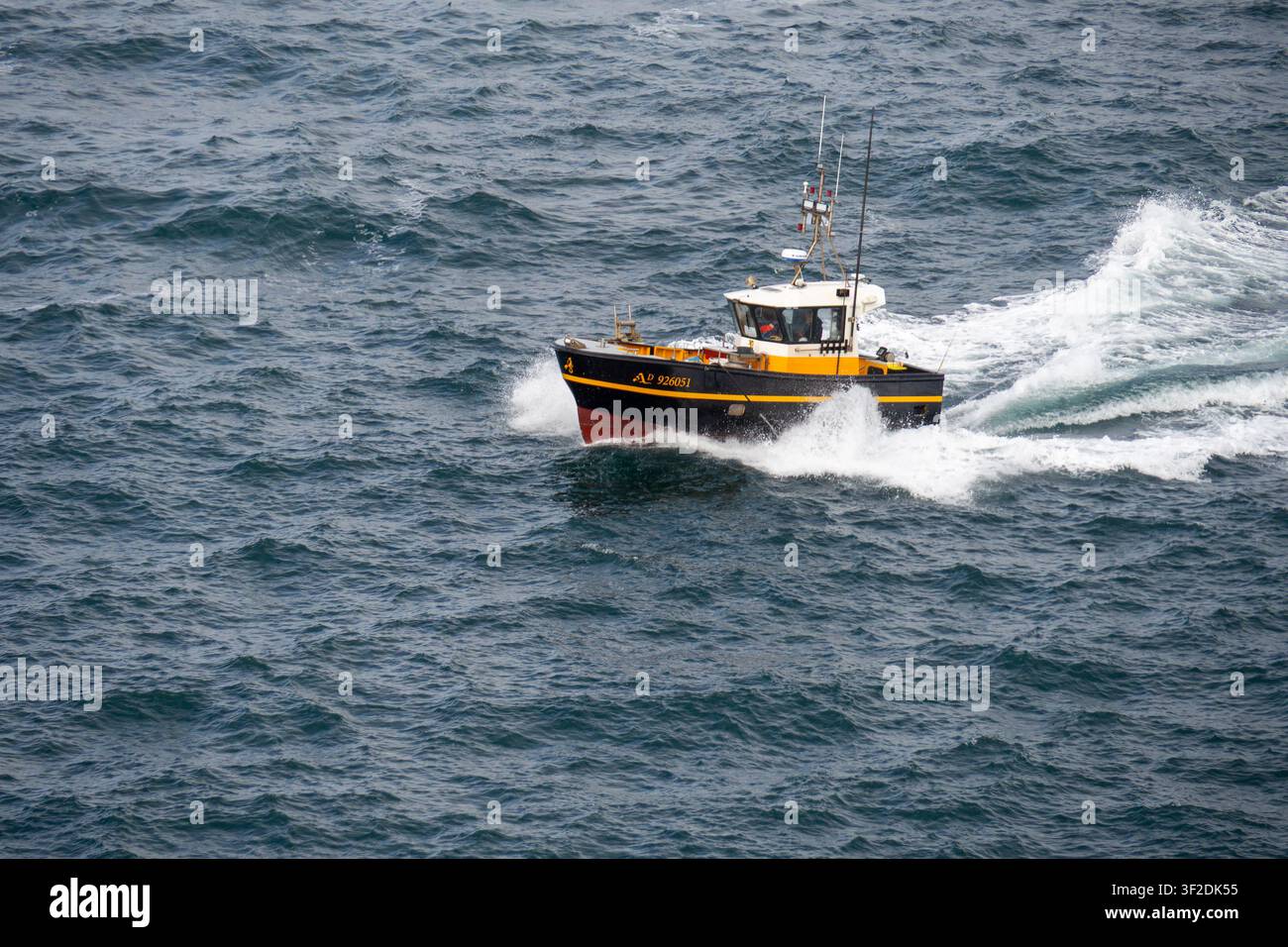 Bateau de pêche jaune et noir fendant une mer agitée bleu foncé avec des éclaboussures blanches et un sillage près d'une côte rocheuse sous un ciel nuageux Banque D'Images