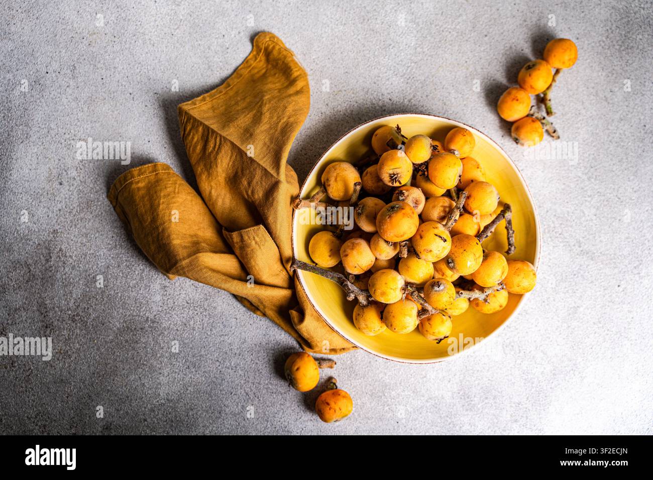 Vue par-dessus de fruits de loquat mûrs dans un bol jaune avec une serviette rustique sur une surface en pierre grise, concept de produits frais sains riches en vitamine C. Banque D'Images