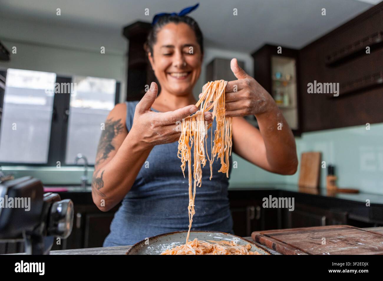 Femme souriante étire des pâtes fraîches faites maison sur une planche farinée dans une cuisine domestique, façonnant des fils pour la cuisson. Préparation de plats de style de vie confortable Banque D'Images
