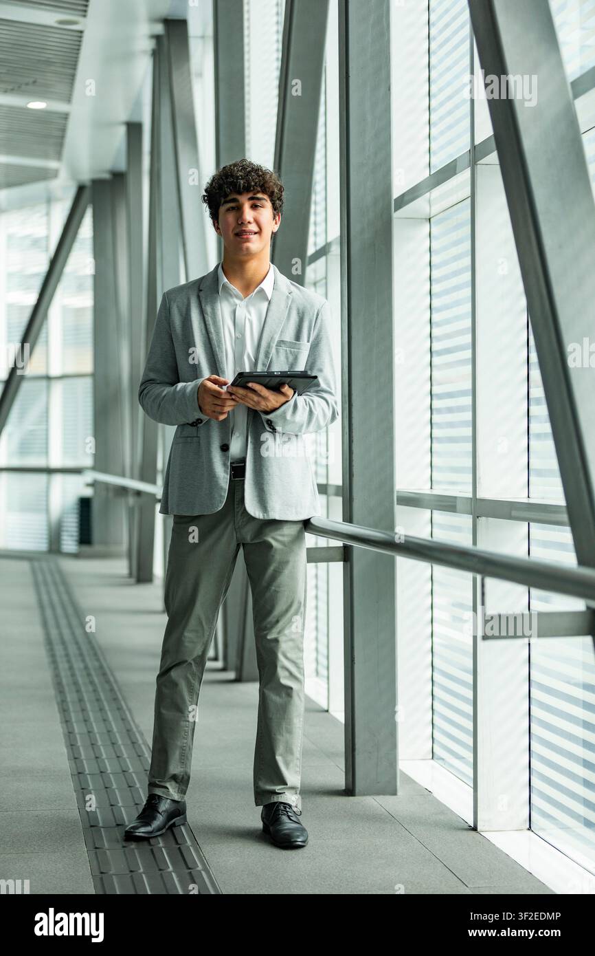 Portrait en pleine longueur d'un jeune professionnel d'affaires en costume léger tenant une tablette dans un couloir vitré moderne et lumineux, suggérant la technologie, fina Banque D'Images