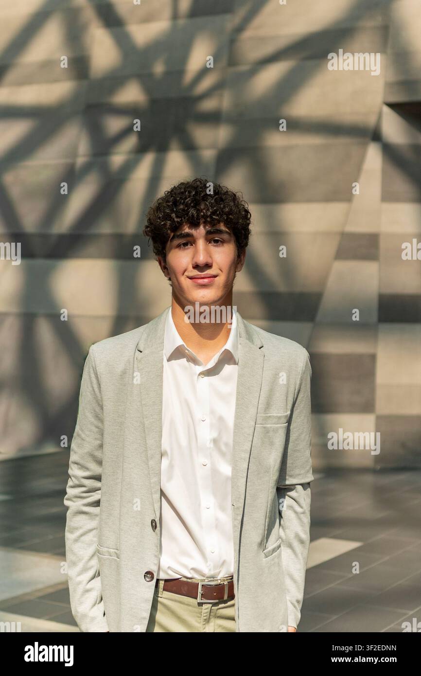 Jeune homme professionnel en costume clair pose à l'extérieur sous un soleil chaud, debout devant un mur géométrique moderne avec des ombres douces, exprimant la confiance Banque D'Images