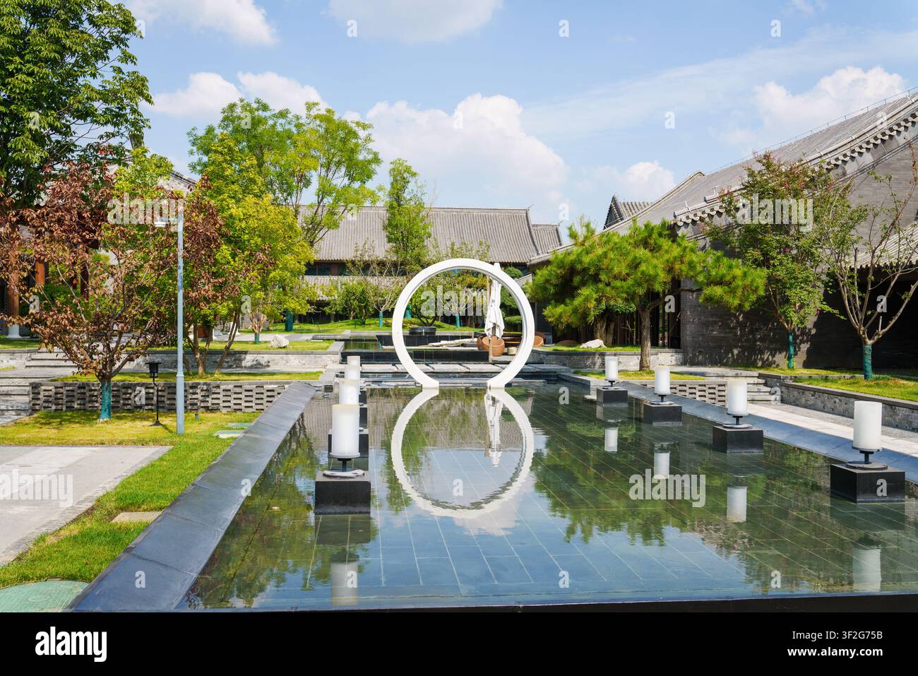 Un long bassin réfléchissant avec une sculpture circulaire blanche traverse un jardin paysager à côté de toits bas de style traditionnel. Banque D'Images