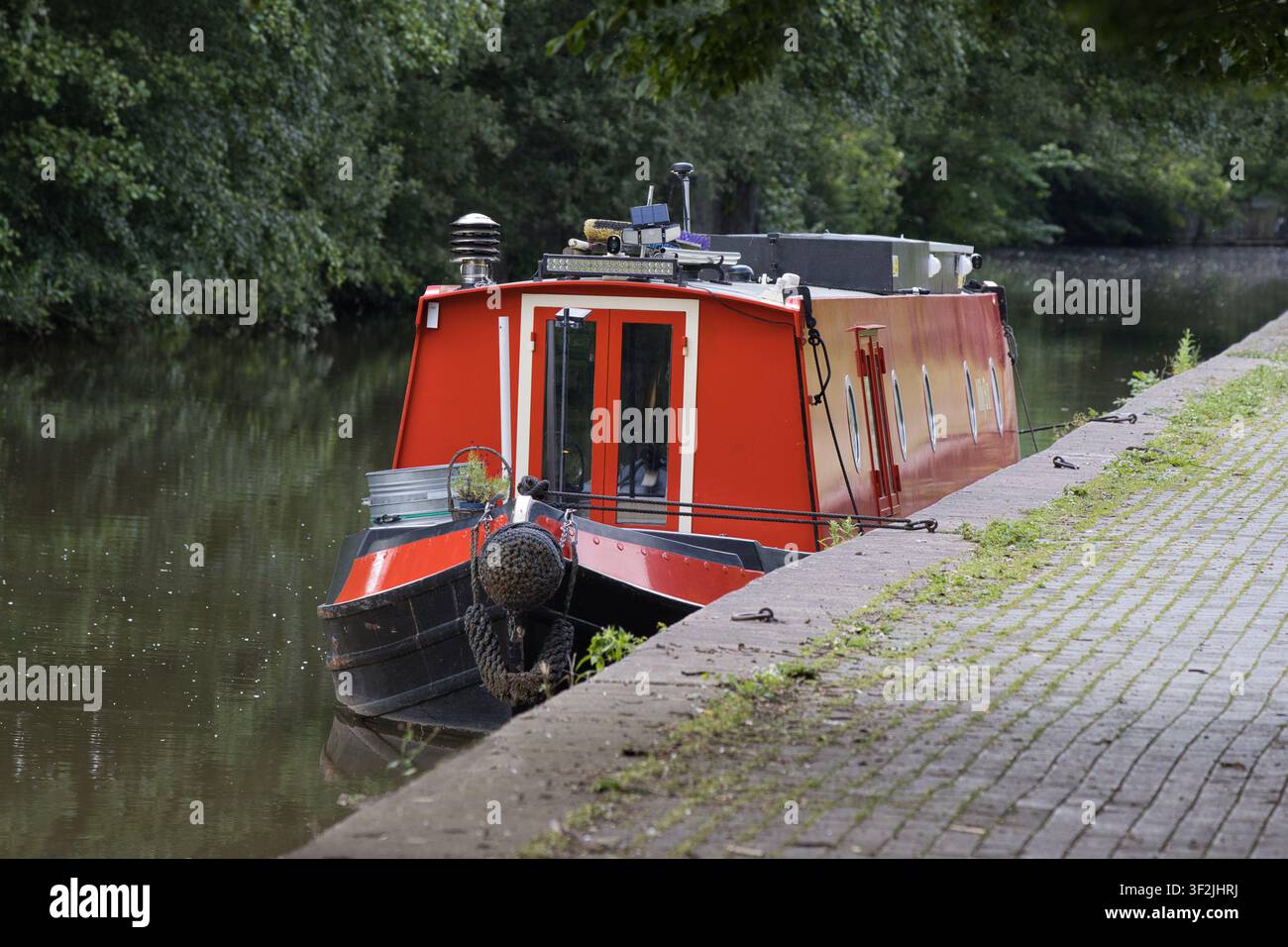Un bateau étroit rouge vif amarré le long d'un chemin de halage en briques sur un canal calme, encadré par des arbres verts denses. Banque D'Images